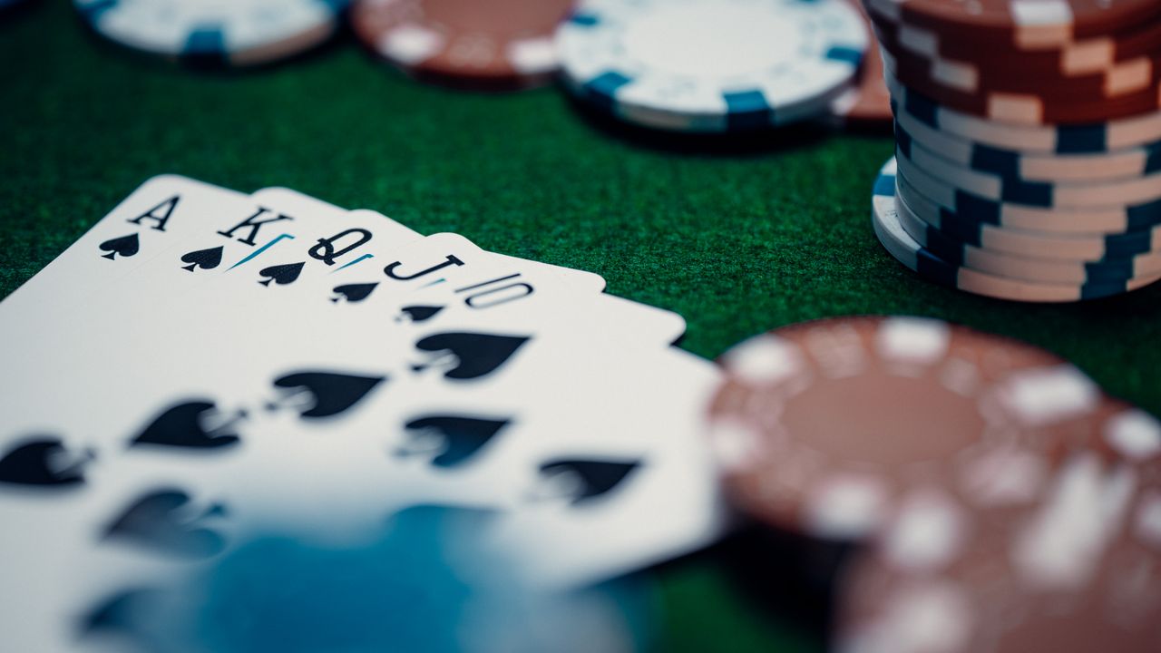 hero-img-01 Close-up of a royal flush poker hand surrounded by colorful chips on a green table.