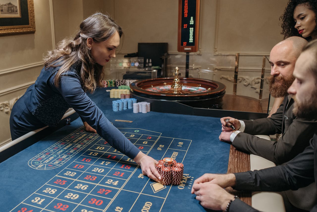 why-choose-us A group of people playing roulette at a casino with a female dealer organizing chips on the table.
